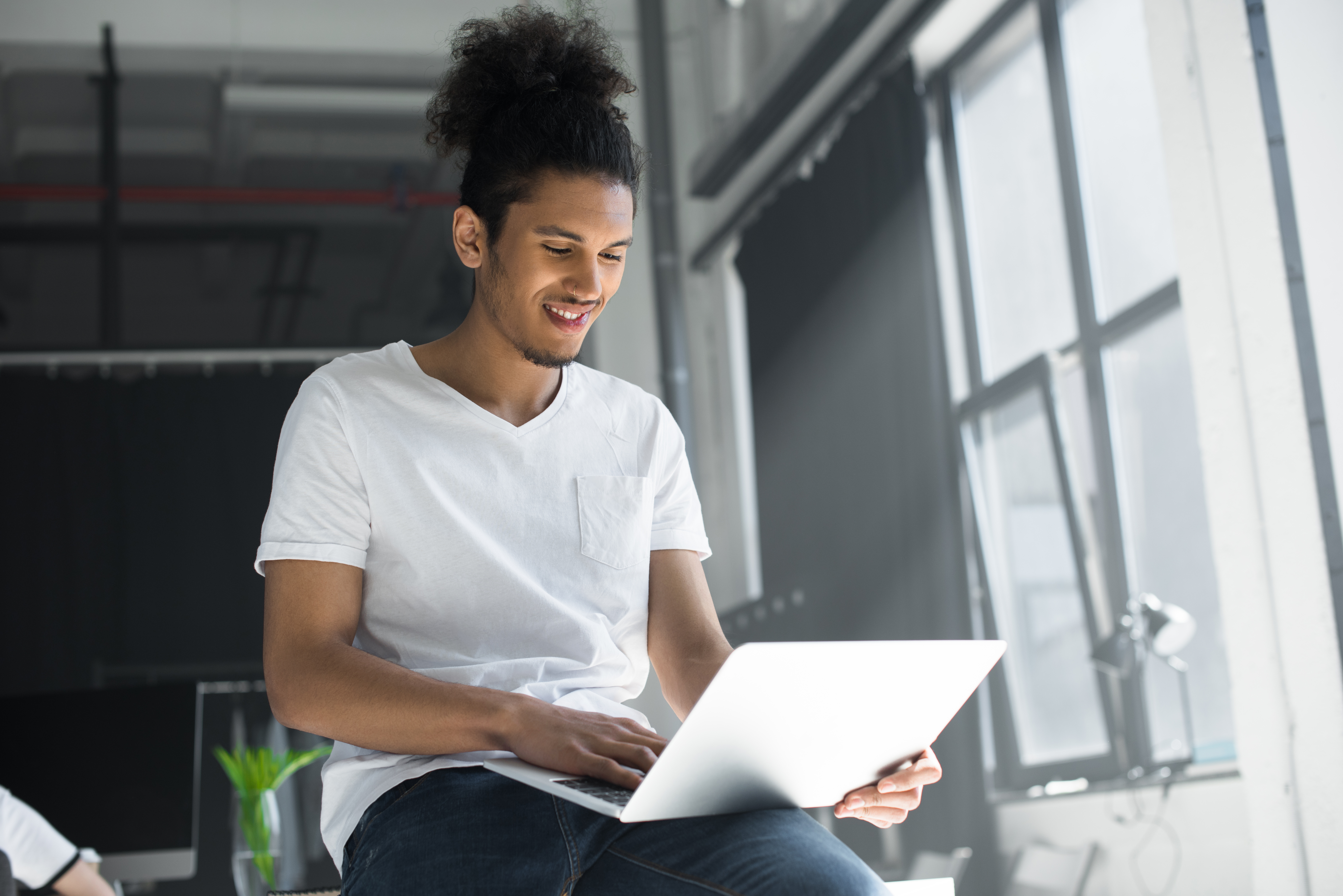 A young man in a white t-shirt and hair in a bun smiles while working on a laptop by a large, sunny window in a modern office, representing flexible online learning.