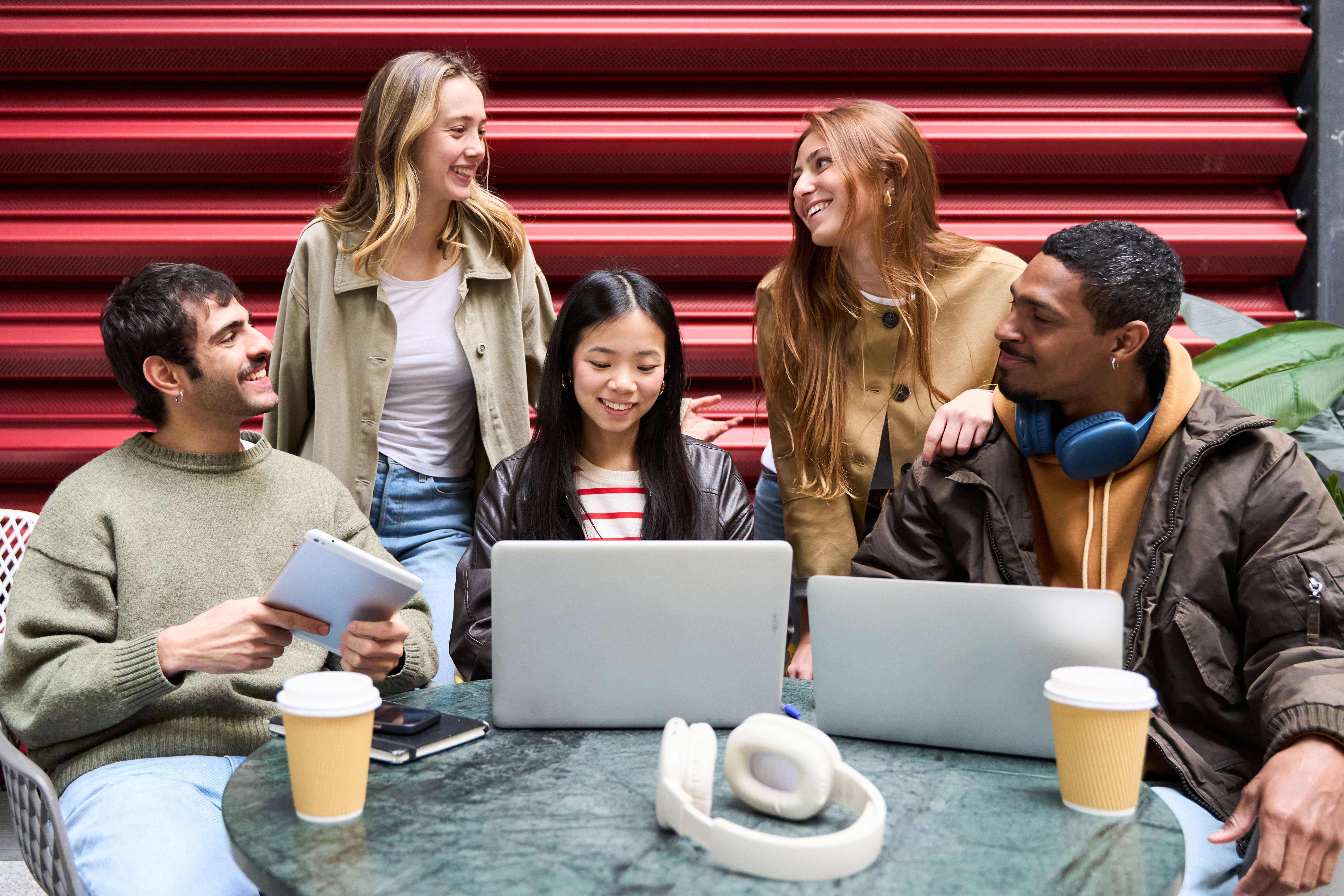 a diverse group of five young people working together on laptops and a tablet at an outdoor table with coffee cups.