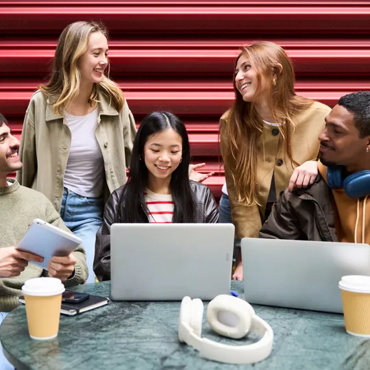 a diverse group of five young people working together on laptops and a tablet at an outdoor table with coffee cups.
