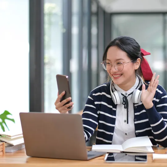 A smiling student wearing glasses and a striped cardigan waves while holding a smartphone for a video call, with a laptop and notebook on a wooden desk for online learning.