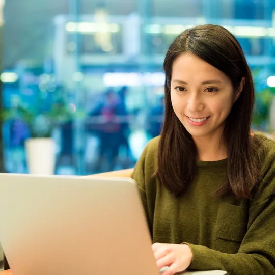 A young woman with curly hair in a black blazer and striped shirt gestures with her hands while speaking to a person opposite her in a modern office, representing a focused computer science discussion or interview.
