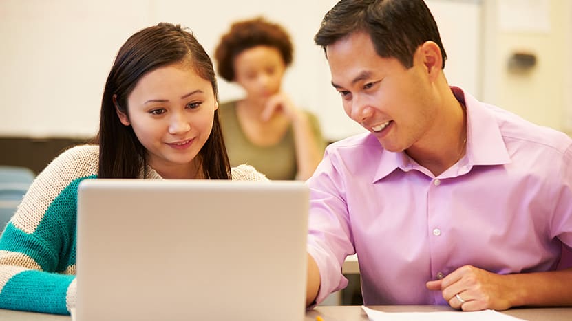 A male teacher or tutor wearing a pink shirt is smiling while leaning in to help a female student look at content on her laptop screen in a classroom setting. Another student is blurred in the background.