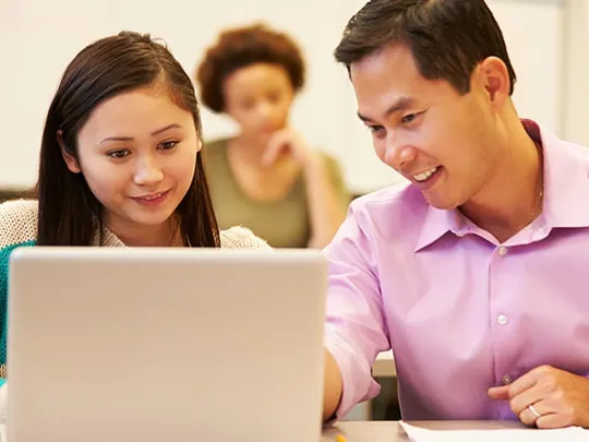 A male teacher or tutor wearing a pink shirt is smiling while leaning in to help a female student look at content on her laptop screen in a classroom setting. Another student is blurred in the background.