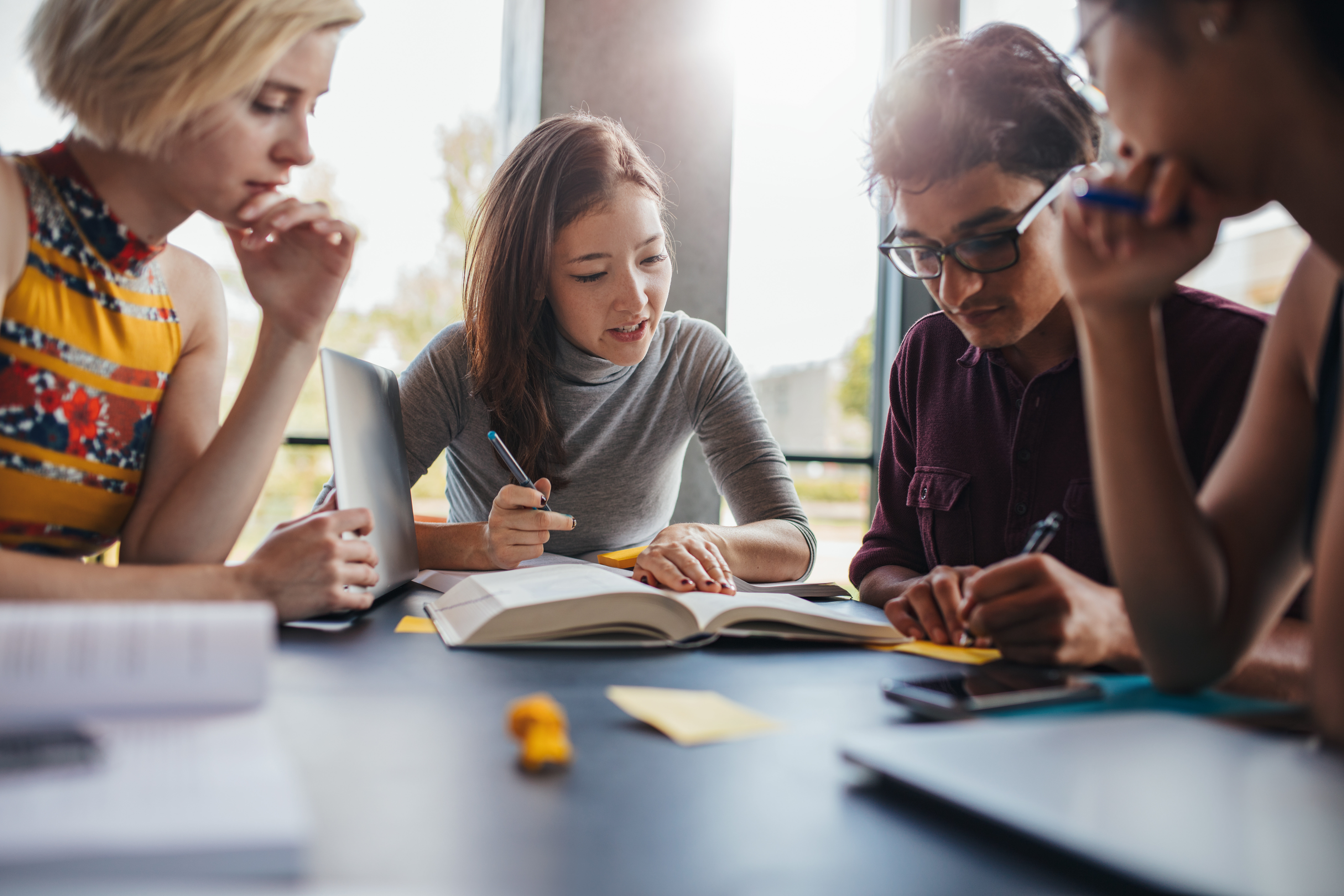 a diverse group of four young students studying, collaborating around a large book and a laptop in a brightly lit room.