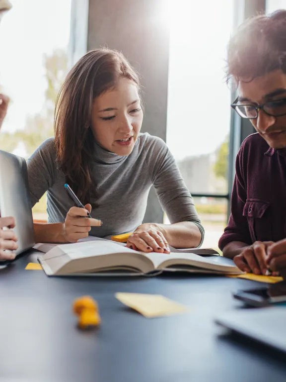 a diverse group of four young students studying, collaborating around a large book and a laptop in a brightly lit room.