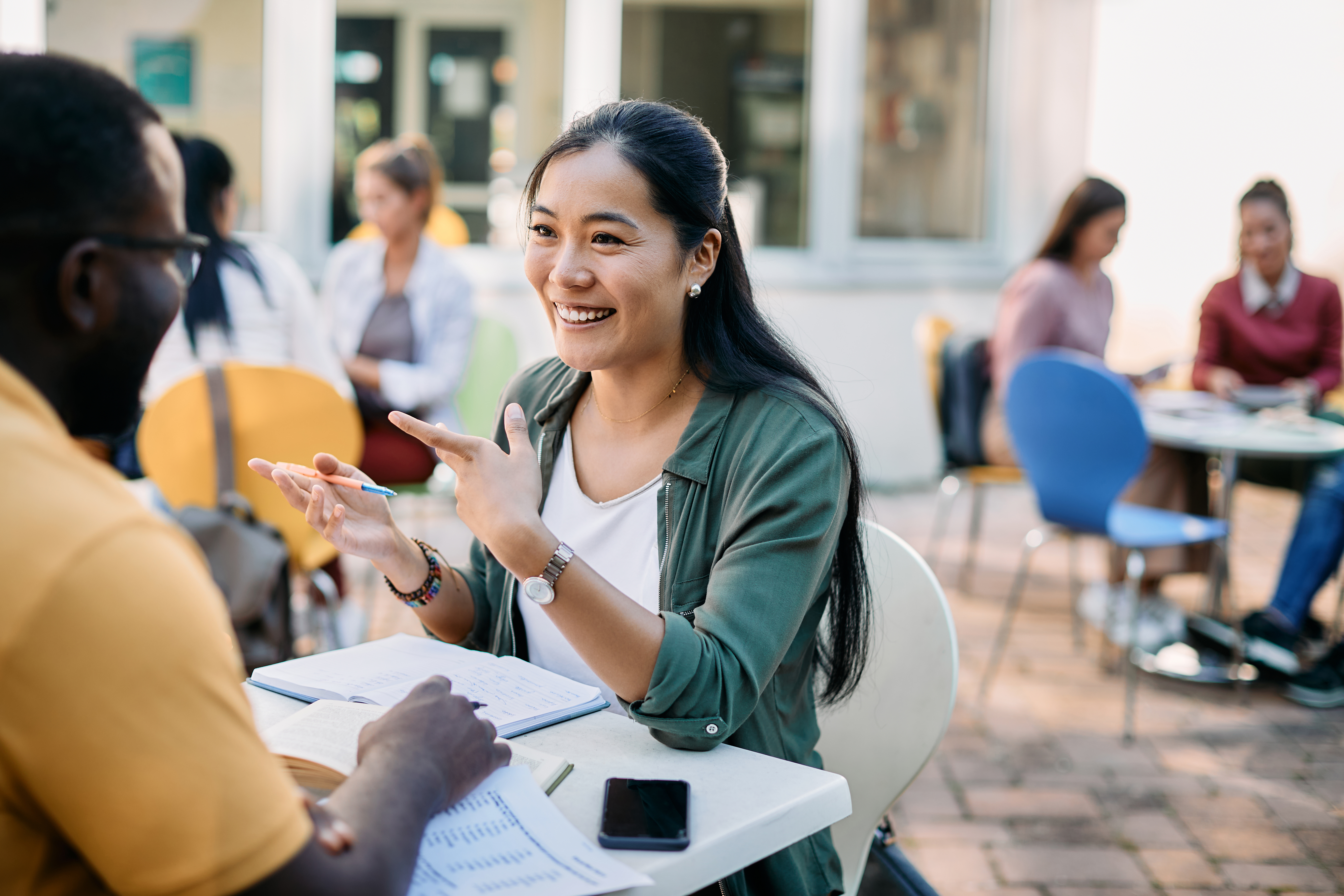 A friendly female student or tutor is sitting across a table from a male student in an outdoor cafe or campus setting, enthusiastically discussing a topic. She is smiling and gesturing with her hands while looking at him. Papers, notebooks, and a smartphone are on the table between them. Other people are visible in the blurred background.