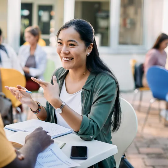 A friendly female student or tutor is sitting across a table from a male student in an outdoor cafe or campus setting, enthusiastically discussing a topic. She is smiling and gesturing with her hands while looking at him. Papers, notebooks, and a smartphone are on the table between them. Other people are visible in the blurred background.