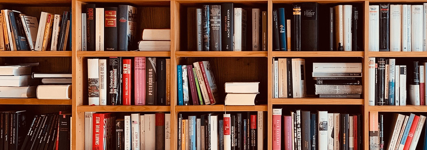 A close-up shot of several wooden bookshelves filled with various books. Some books are standing upright, while others are stacked horizontally. The spines of the books are different colors and have different titles, although they are not clearly readable.