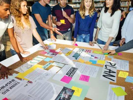 A diverse group of college students collaborating on a project, gathering around a table covered with papers, plans, and sticky notes.