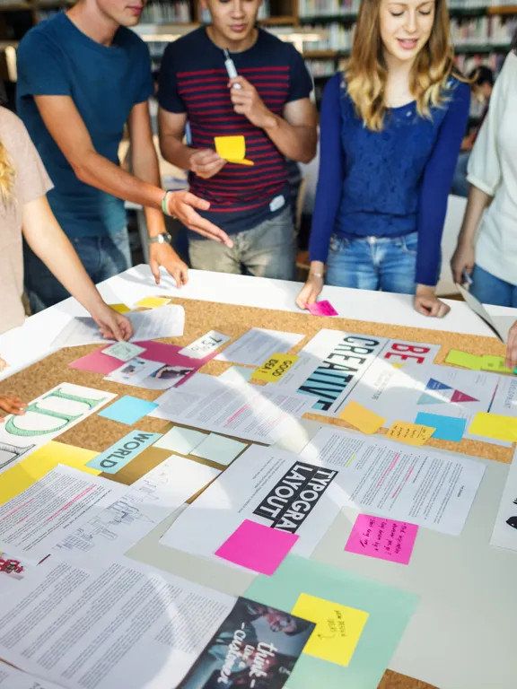 A diverse group of college students collaborating on a project, gathering around a table covered with papers, plans, and sticky notes.