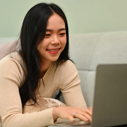 A young man with curly hair and a black sweater smiles while talking to another person in a lively, plant-filled cafe, representing an academic discussion.