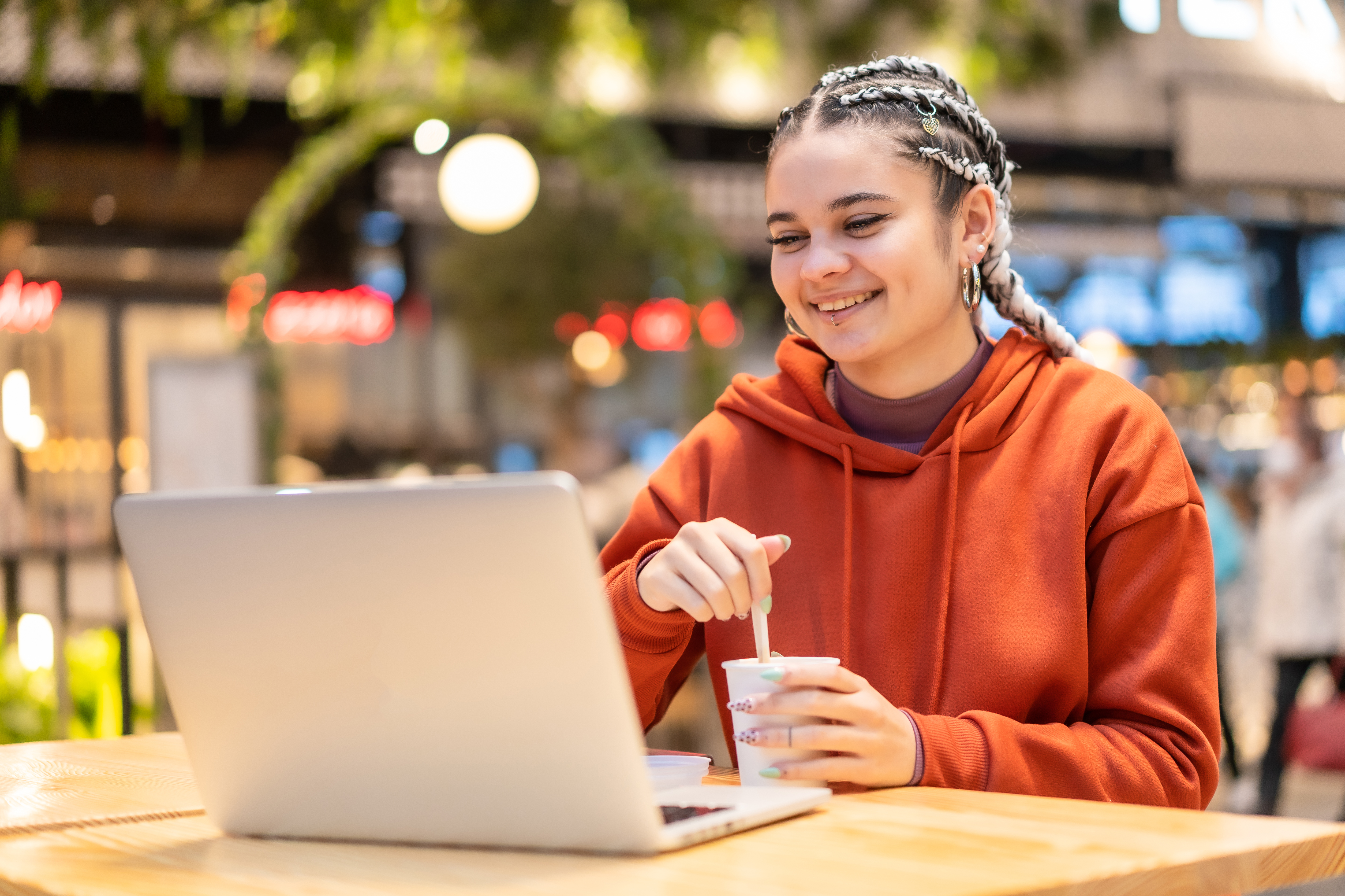 A smiling female student with braided hair, wearing an orange hoodie, using a laptop and stirring a drink while studying in a brightly lit indoor/outdoor cafe area.