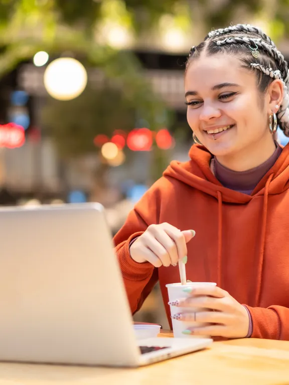 A smiling female student with braided hair, wearing an orange hoodie, using a laptop and stirring a drink while studying in a brightly lit indoor/outdoor cafe area.