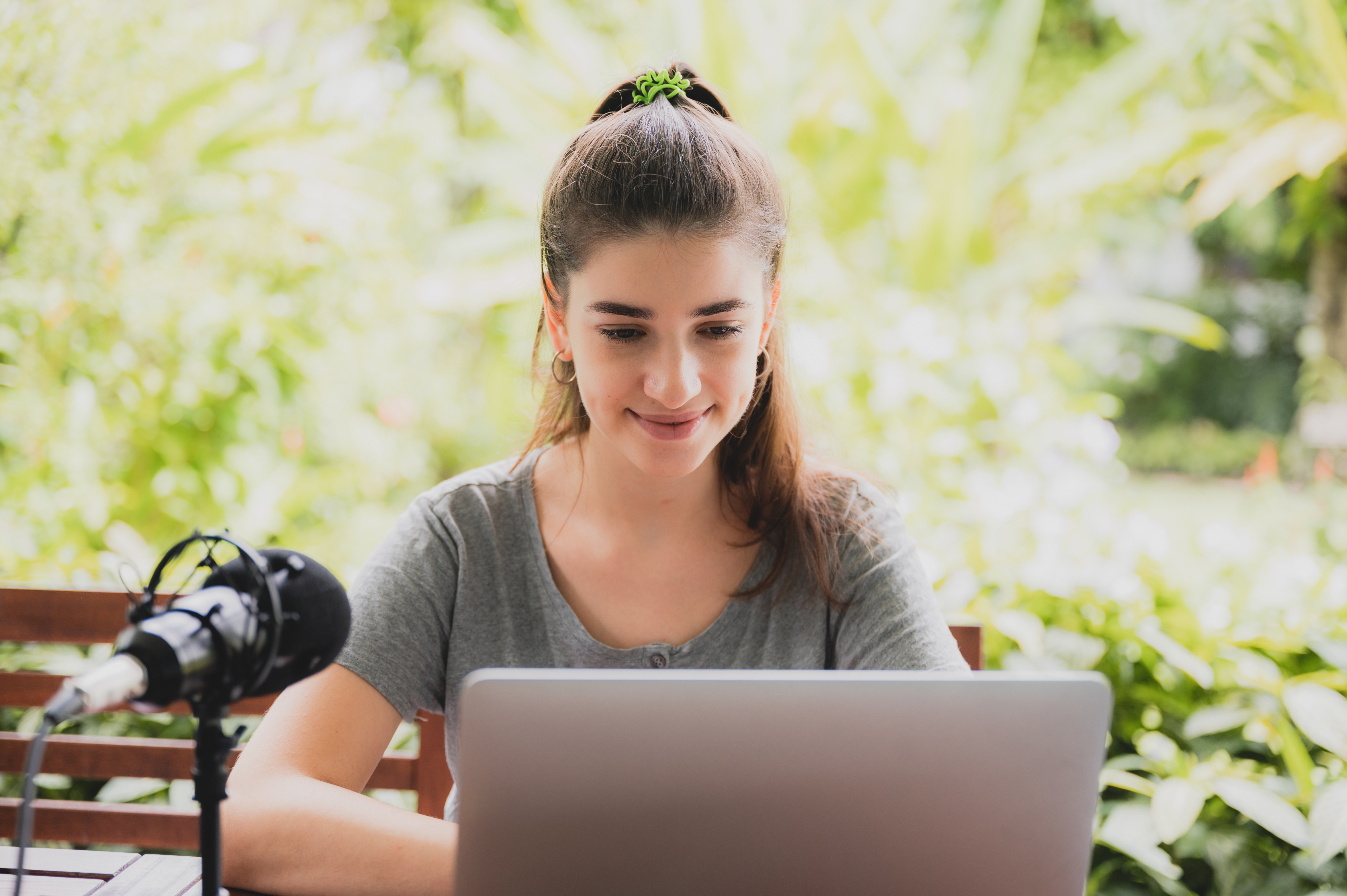 A young woman with her hair in a ponytail smiles while working on a laptop with a professional microphone in an outdoor, lush green setting, representing flexible online learning.