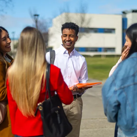 A diverse group of four smiling students on a college campus, one man holding an orange folder, engaged in conversation outdoors on a sunny day.