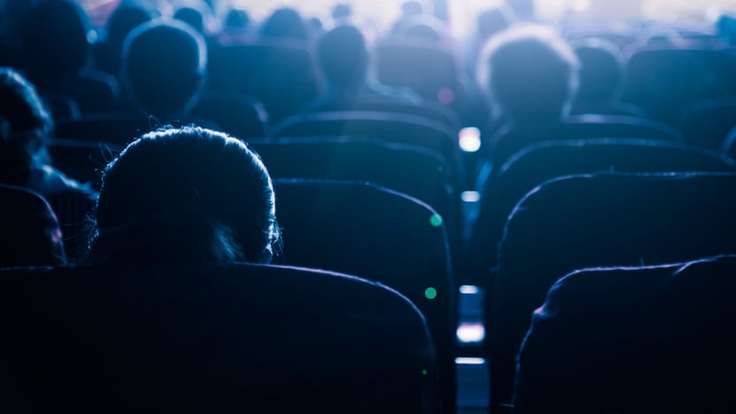  A view from behind of people sitting in a dark cinema or theater, watching a screen. The light from the screen illuminates the tops of their heads and the seats.