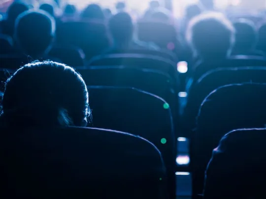 A view from behind of people sitting in a dark cinema or theater, watching a screen. The light from the screen illuminates the tops of their heads and the seats.