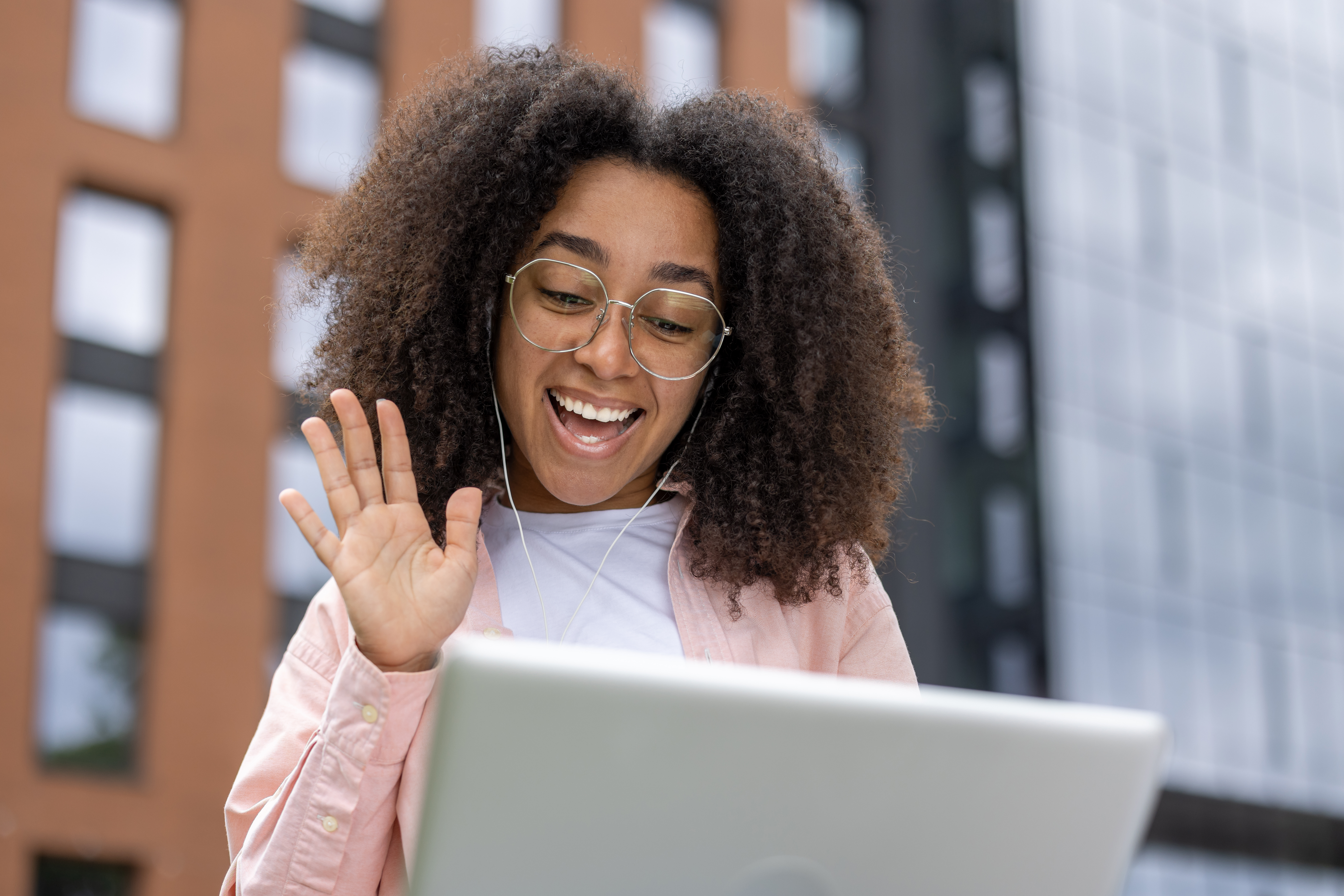 A young woman with curly hair and glasses smiles and waves during an outdoor video call on her laptop, representing flexible online learning.