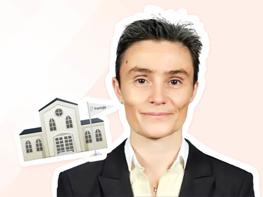 Professional headshot of a smiling person wearing a black suit jacket and white shirt, with a short, dark haircut. Next to them is a small sticker illustration of a school building with a flag displaying the word 'Pamoja'.