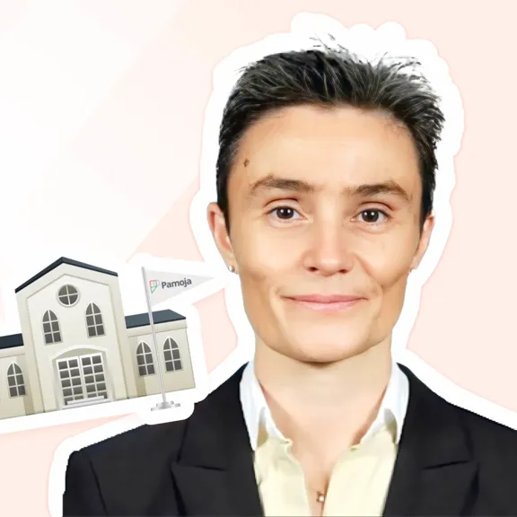 Professional headshot of a smiling person wearing a black suit jacket and white shirt, with a short, dark haircut. Next to them is a small sticker illustration of a school building with a flag displaying the word 'Pamoja'.