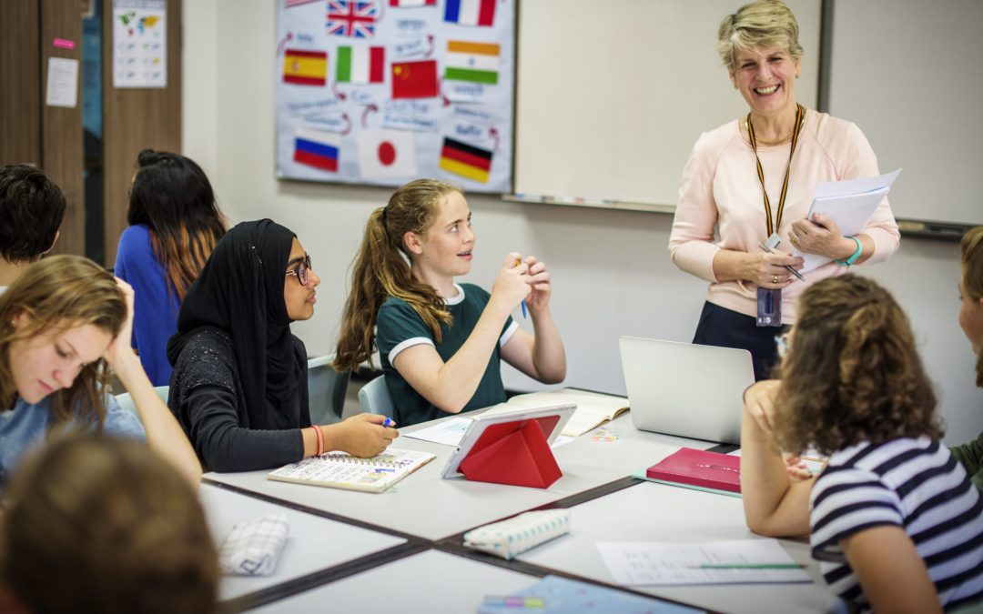 A diverse group of students, including a female student in a black hijab, are seated around a table, actively engaged in a class discussion. A smiling female teacher stands at the whiteboard, holding papers, with a language-themed poster of world flags behind her.