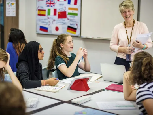 A diverse group of students, including a female student in a black hijab, are seated around a table, actively engaged in a class discussion. A smiling female teacher stands at the whiteboard, holding papers, with a language-themed poster of world flags behind her.