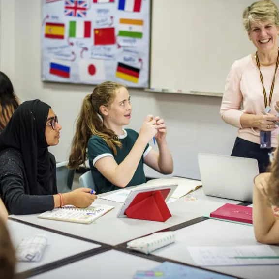 A diverse group of students, including a female student in a black hijab, are seated around a table, actively engaged in a class discussion. A smiling female teacher stands at the whiteboard, holding papers, with a language-themed poster of world flags behind her.