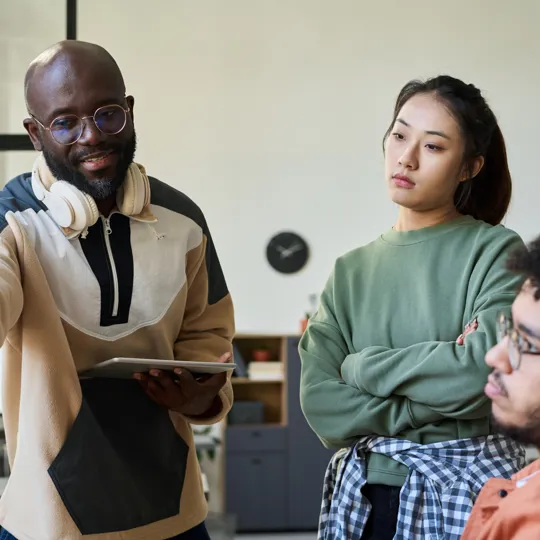Two students, a young woman in a yellow sweater and a young man in a green sweatshirt, collaborate on a whiteboard with markers in a bright classroom.