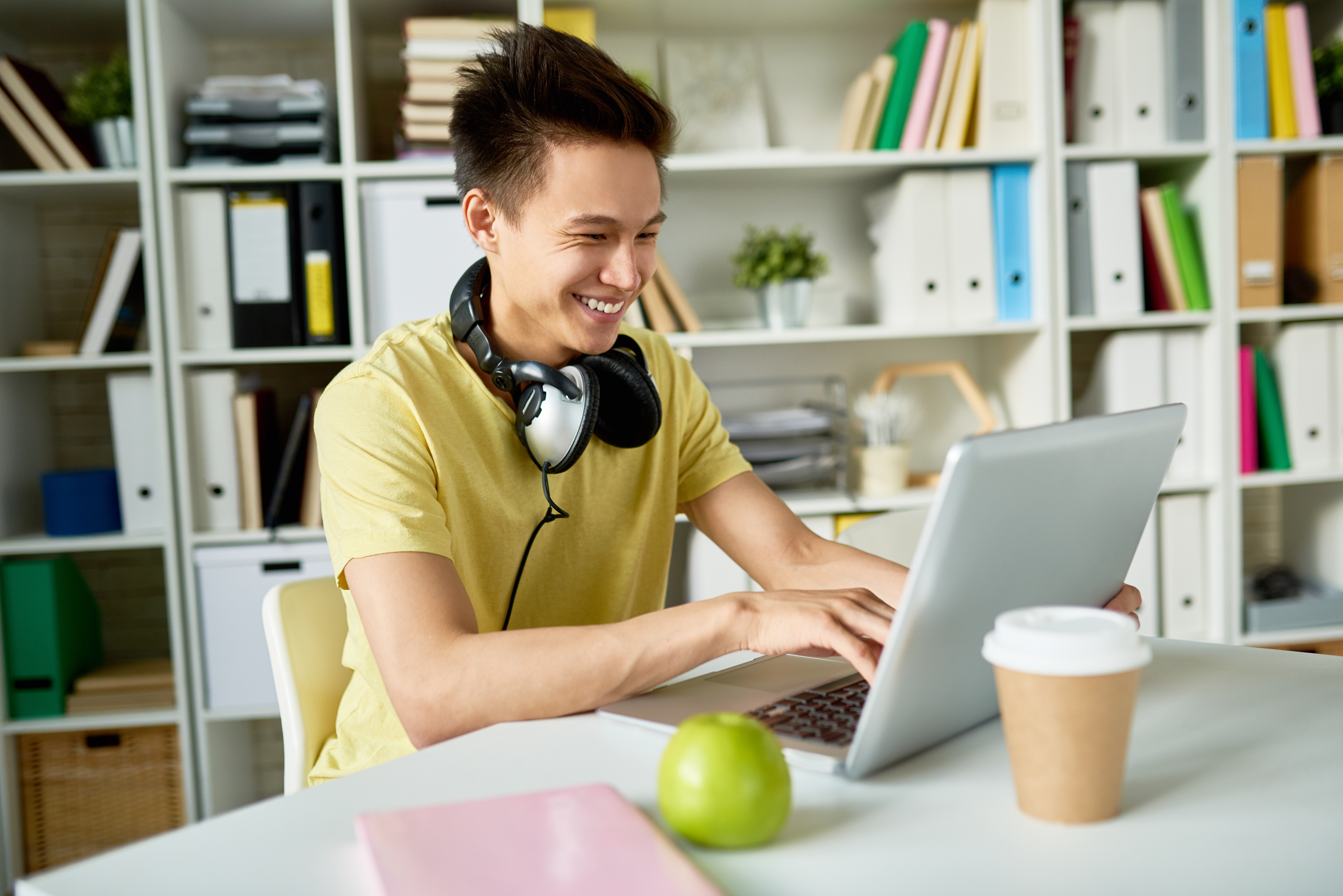 A smiling young man in a yellow shirt and headphones works on a laptop at a bright office desk with an apple and coffee, representing online learning for philosophy.