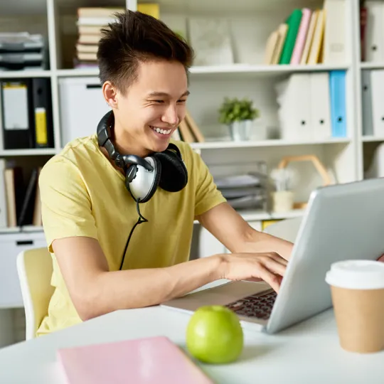 A smiling young man in a yellow shirt and headphones works on a laptop at a bright office desk with an apple and coffee, representing online learning for philosophy.