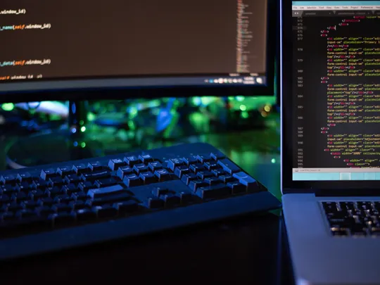 A young woman in a green sweater and glasses smiles while focused on working on a silver laptop in a brightly lit, modern indoor public area.