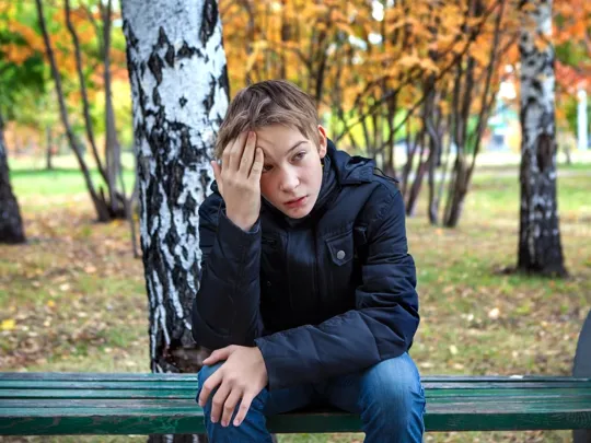 Young boy sat outside on a bench with his hand on his head.