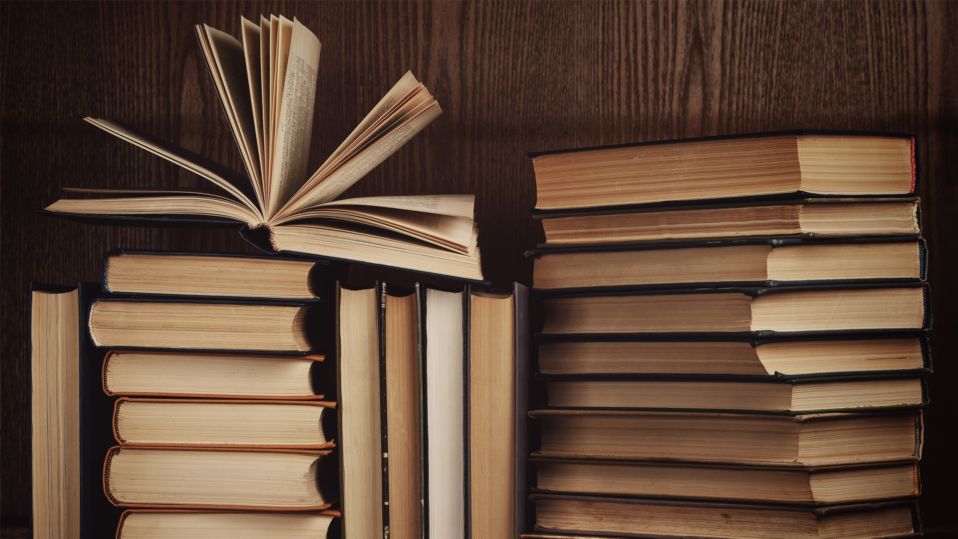 A close-up of a stack of old books with warm-toned pages, one open and fanned out, set against a dark wooden bookshelf.