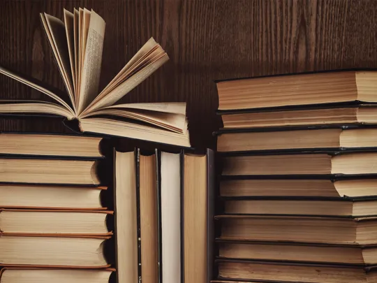 A close-up of a stack of old books with warm-toned pages, one open and fanned out, set against a dark wooden bookshelf.
