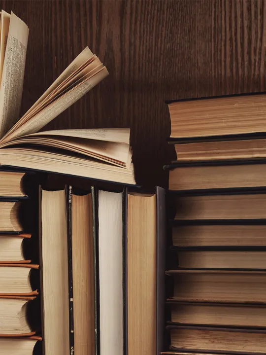 A close-up of a stack of old books with warm-toned pages, one open and fanned out, set against a dark wooden bookshelf.