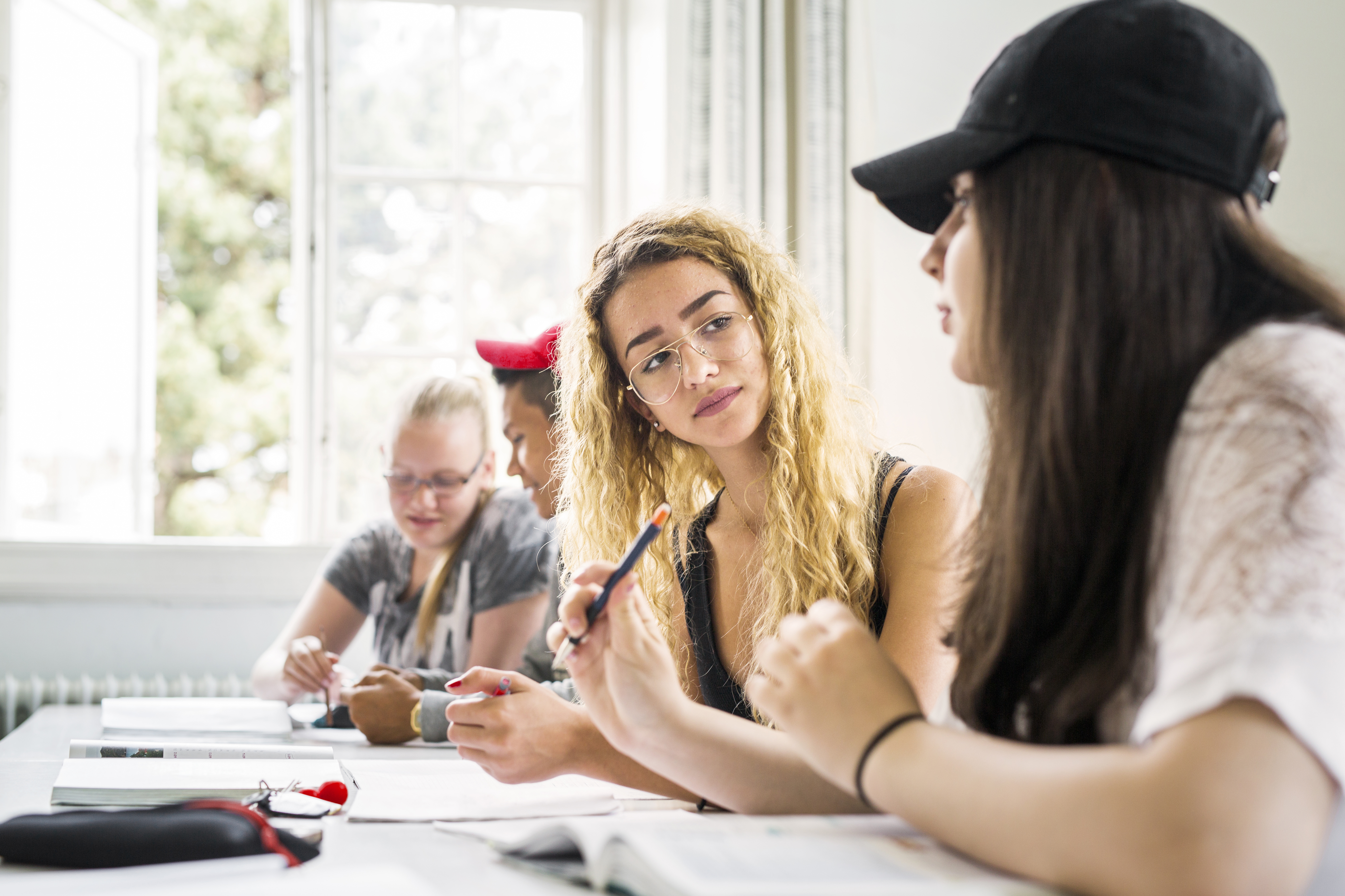 Two female students with long hair discussing a lesson in a classroom, with other students working at tables behind them.