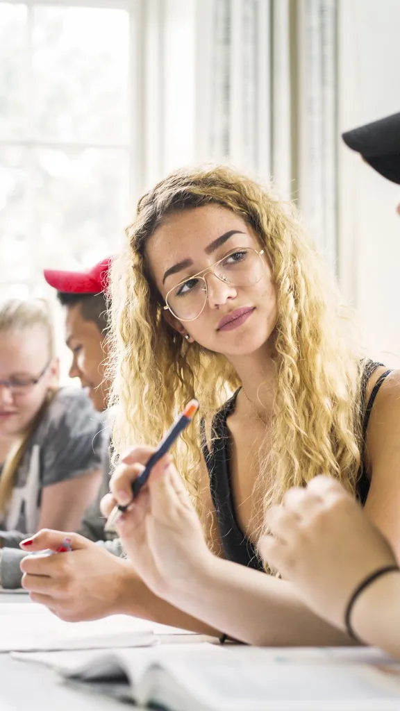 Two female students with long hair discussing a lesson in a classroom, with other students working at tables behind them.