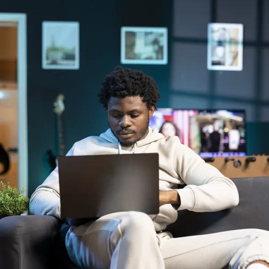 A young man in a light hoodie sits on a couch in a dimly lit, modern living room, working on a black laptop.