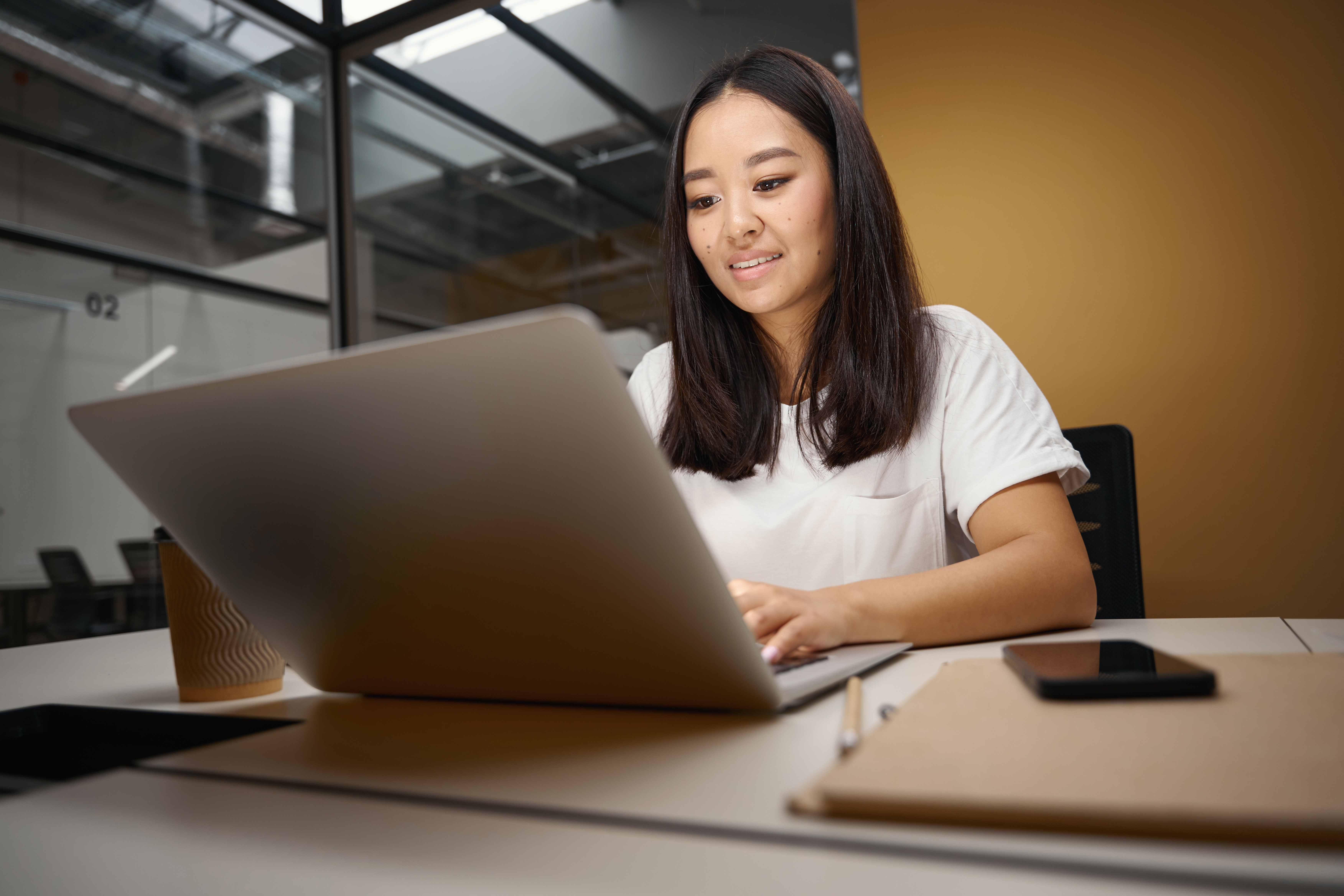 A young Asian woman in a white t-shirt smiles while working on a laptop at a bright office desk, representing flexible online learning.