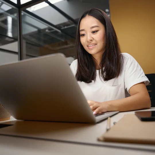 A young Asian woman in a white t-shirt smiles while working on a laptop at a bright office desk, representing flexible online learning.