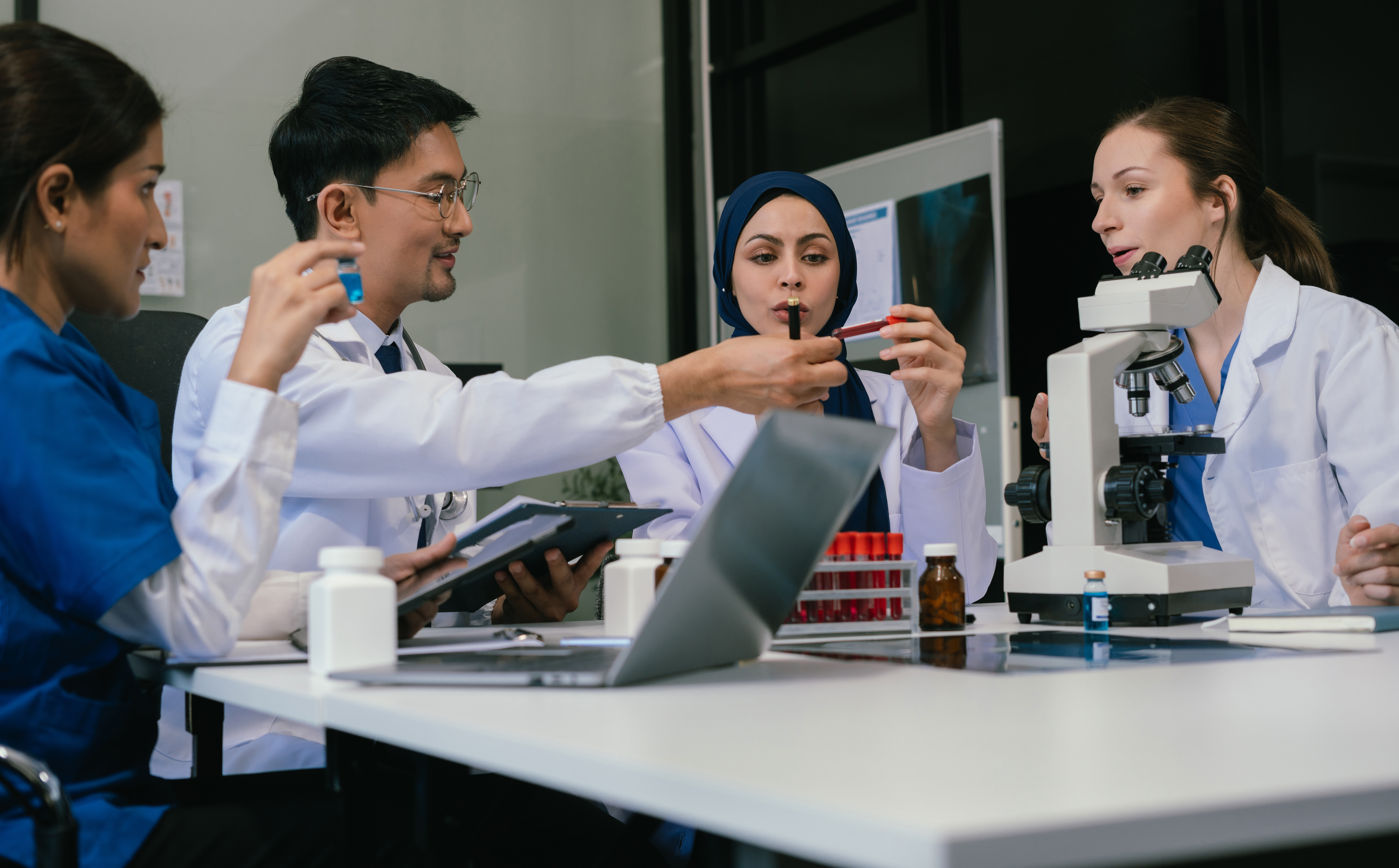 A team of four diverse scientists and medical professionals collaborating at a table with a microscope, test tubes, and a laptop, in a brightly lit laboratory setting.