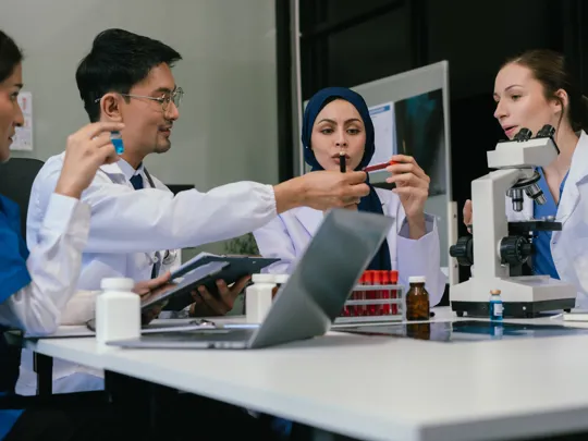 A team of four diverse scientists and medical professionals collaborating at a table with a microscope, test tubes, and a laptop, in a brightly lit laboratory setting.