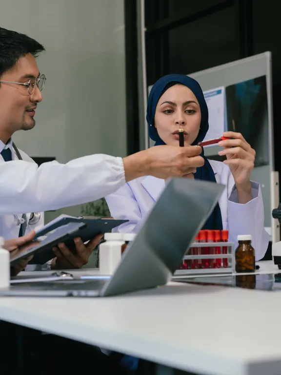 A team of four diverse scientists and medical professionals collaborating at a table with a microscope, test tubes, and a laptop, in a brightly lit laboratory setting.