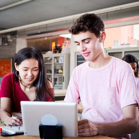 Three adults collaborate in a modern office, with a man pointing at a computer screen while holding a tablet, representing digital society group work.