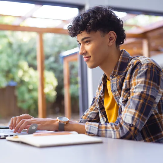 A young student with curly hair smiles while working on a laptop at an outdoor table under a patio cover, with green trees in the background.