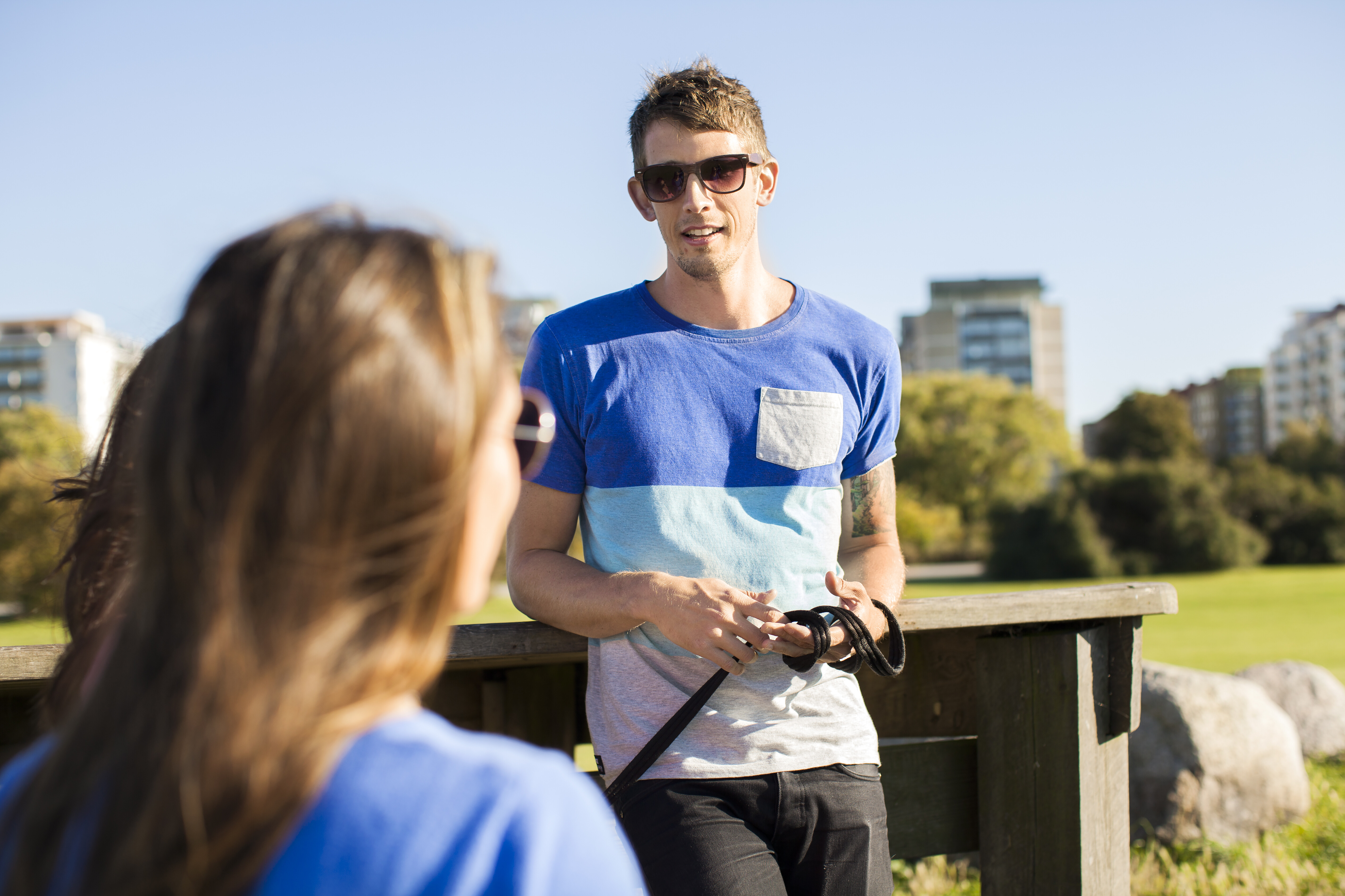 A young man wearing sunglasses and a blue and white striped t-shirt stands outside in a park, leaning on a wooden railing and talking to a person whose back is to the camera. Apartment buildings are visible in the blurred background.