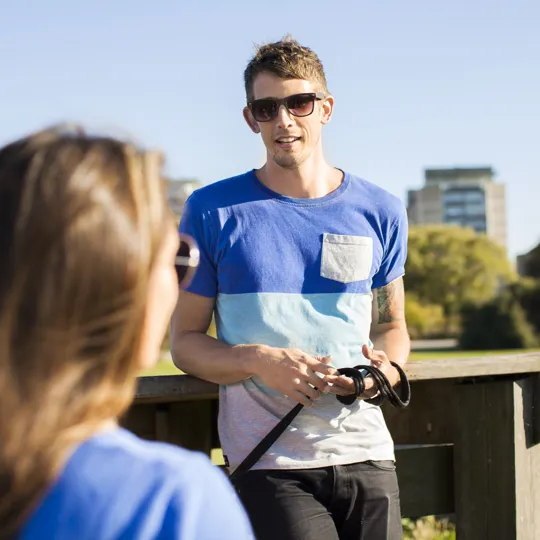A young man wearing sunglasses and a blue and white striped t-shirt stands outside in a park, leaning on a wooden railing and talking to a person whose back is to the camera. Apartment buildings are visible in the blurred background.