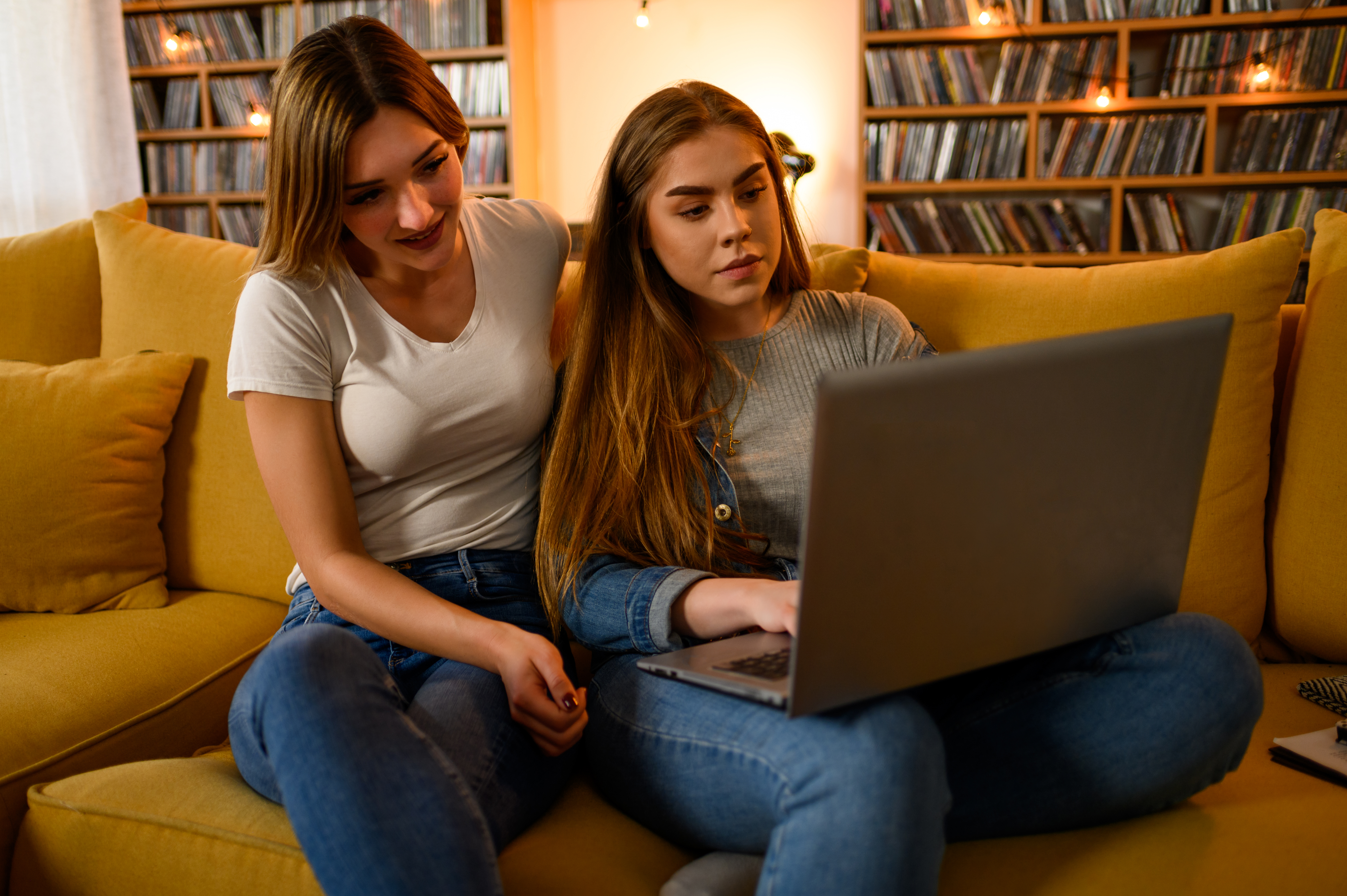 a young woman sitting next to a teenage girl on a yellow couch, looking at a laptop together with a large shelf of media in the background.