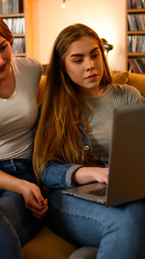 a young woman sitting next to a teenage girl on a yellow couch, looking at a laptop together with a large shelf of media in the background.