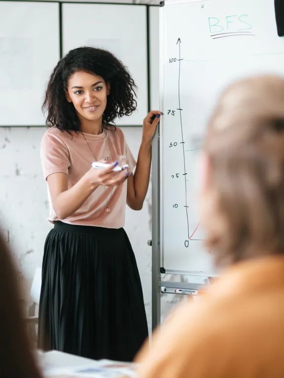 A female teacher with curly dark hair smiles while presenting a graph on a whiteboard to a group of seated students in a modern classroom.